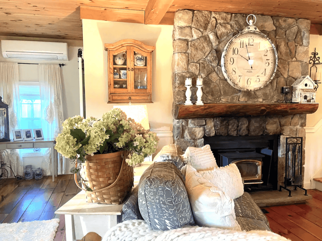 Cozy living room with a stone fireplace, large wall clock, wicker basket of hydrangeas, and plush sofas. decor includes a wooden display cabinet and curtains. Short term rental New Hampshire
