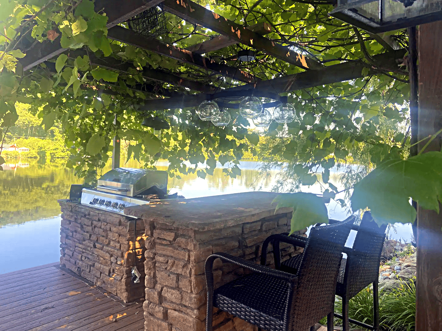 Outdoor kitchen setup on a lakeside wooden deck with a stone counter, grill, seating under a leafy pergola, ideal for short-term rental in New Hampshire.